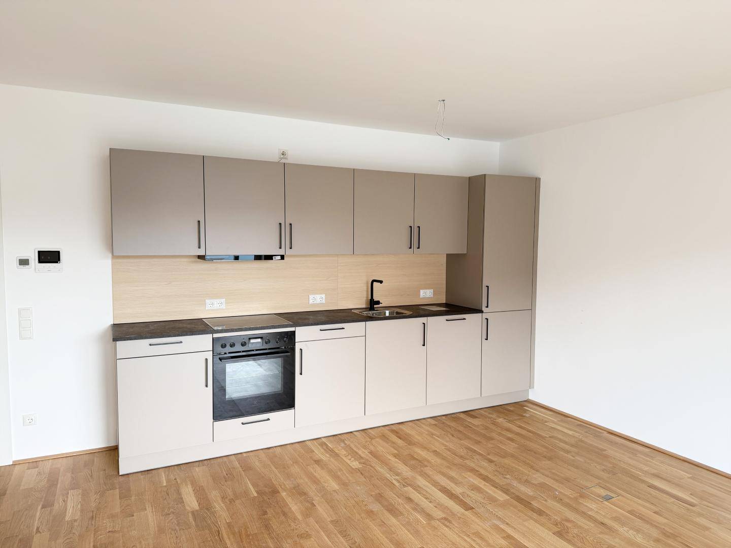 Moderne Küchenzeile mit beigen Schränken, dunkler Arbeitsplatte und integriertem Backofen in offenem Wohnbereich.