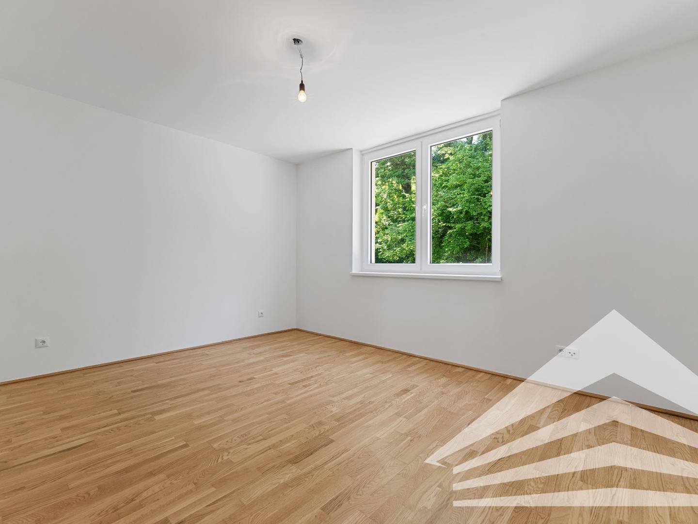 Helles, leeres Schlafzimmer mit Holzboden und großem Fenster mit Grünblick.