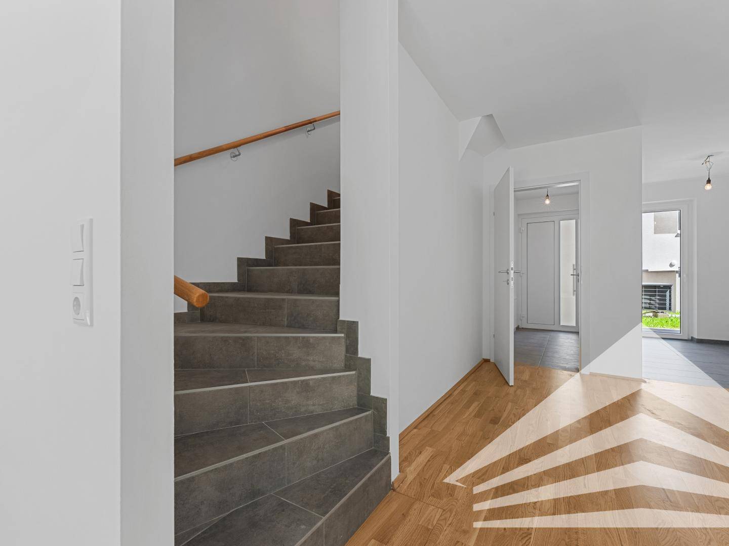 Treppenhaus mit grauen Fliesenstufen und Holzgeländer, führt zu einem hellen Flurbereich.
