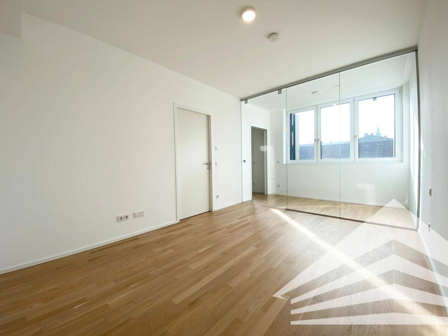 Heller Raum mit Holzboden und großen Fenstern, die viel Licht hereinlassen.
