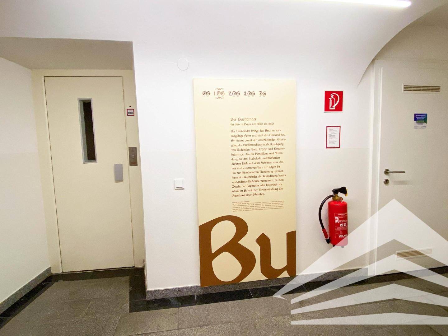 Heller Flur mit Aufzug und Informationstafel, ausgestattet mit einem Feuerlöscher für Sicherheit.