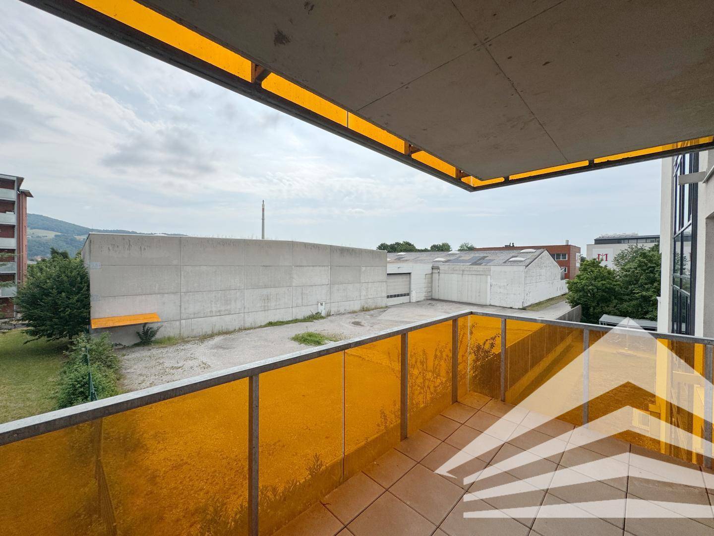 Balkon mit orangefarbener Brüstung und Blick auf angrenzende Gebäude und eine offene Fläche.