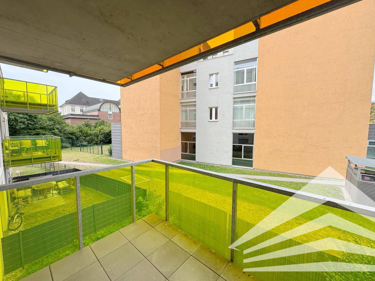Heller Balkon mit gelb-grüner Brüstung und Ausblick auf die benachbarten Gebäude und eine Rasenfläche.