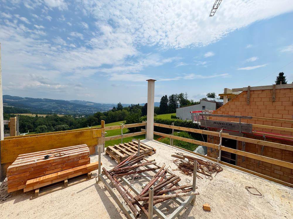 Baustelle mit Betonboden und Baumaterialien, umgeben von einer weiten grünen Hügellandschaft.