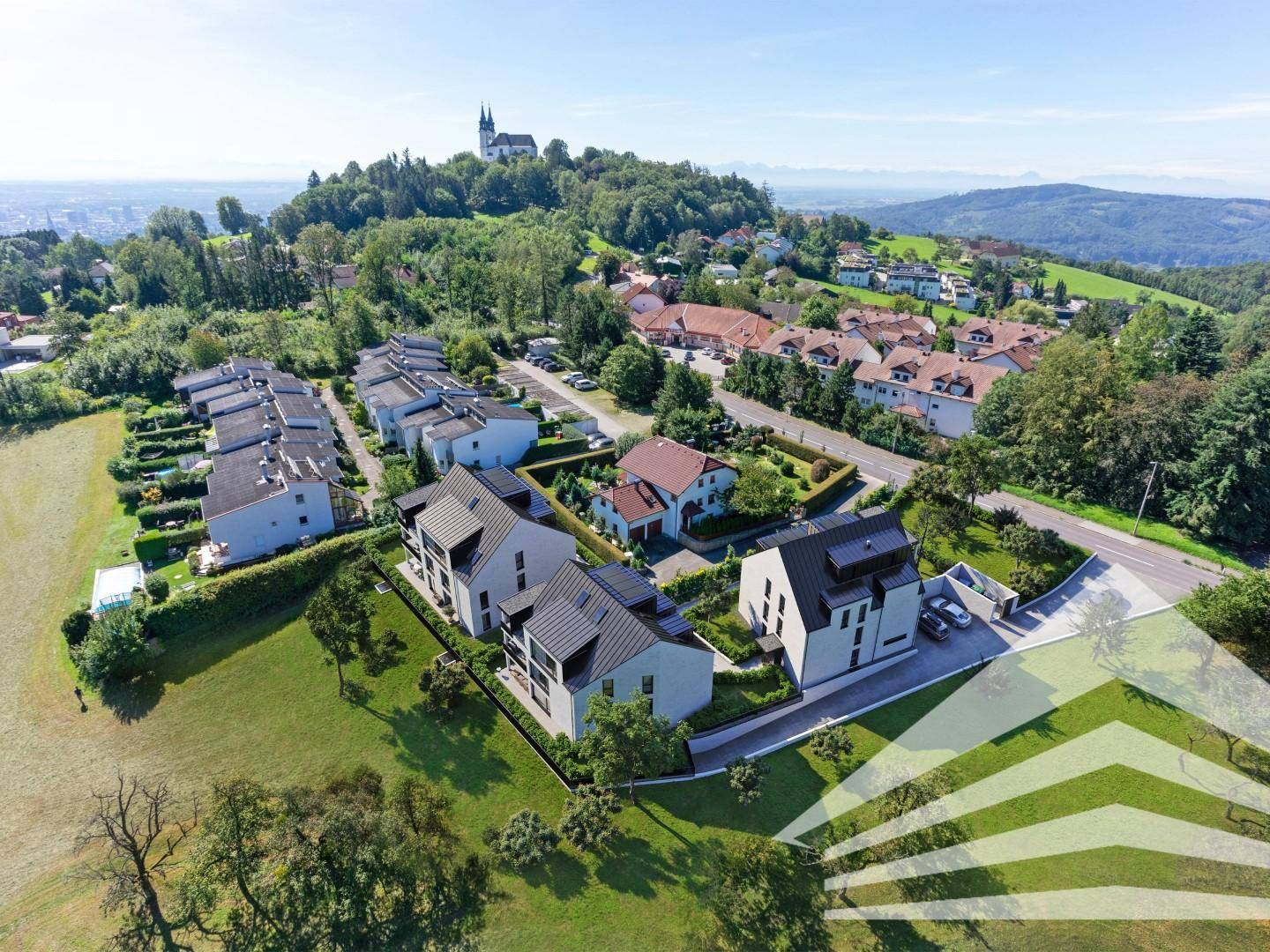 Luftaufnahme einer Wohnsiedlung mit Neubauten und viel Grün, Kirche auf dem Hügel im Hintergrund.