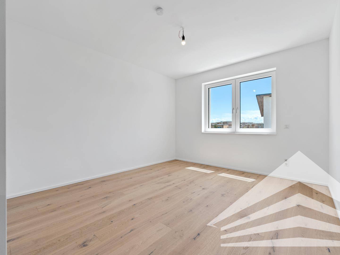 Helles, leeres Schlafzimmer mit Holzboden und großem Fenster, das viel Tageslicht hereinlässt.