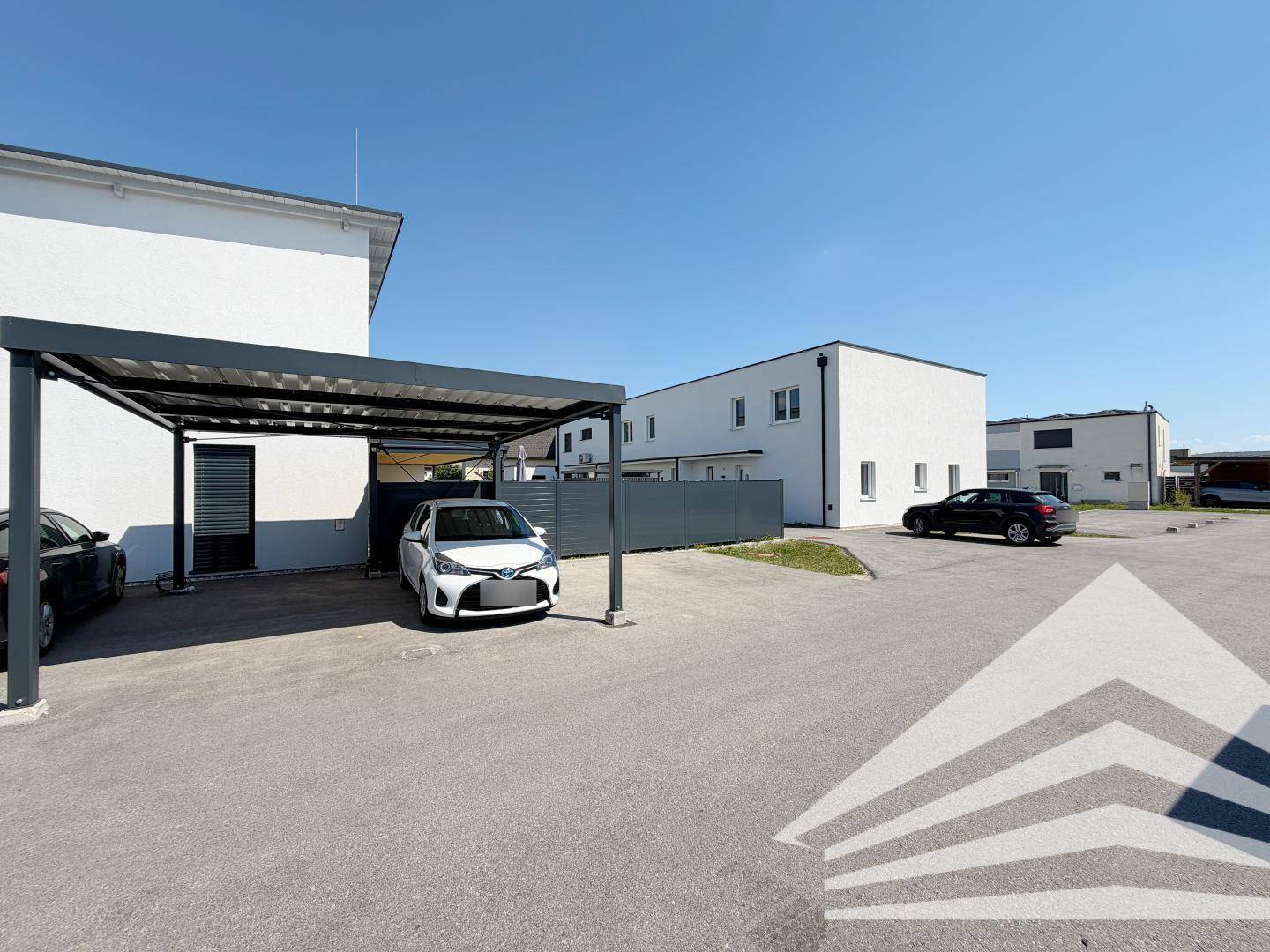 Parkplatz mit Carport vor einem modernen Wohngebäude unter blauem Himmel.