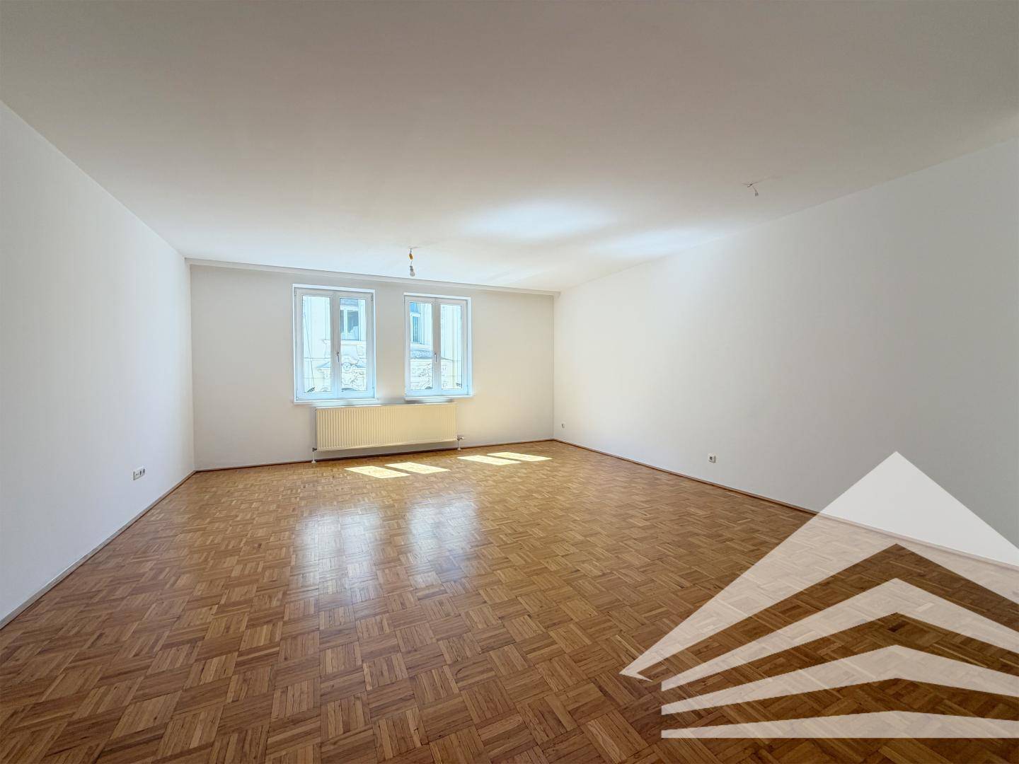 Geräumiges, helles Wohnzimmer mit Parkettboden und zwei großen Fenstern.