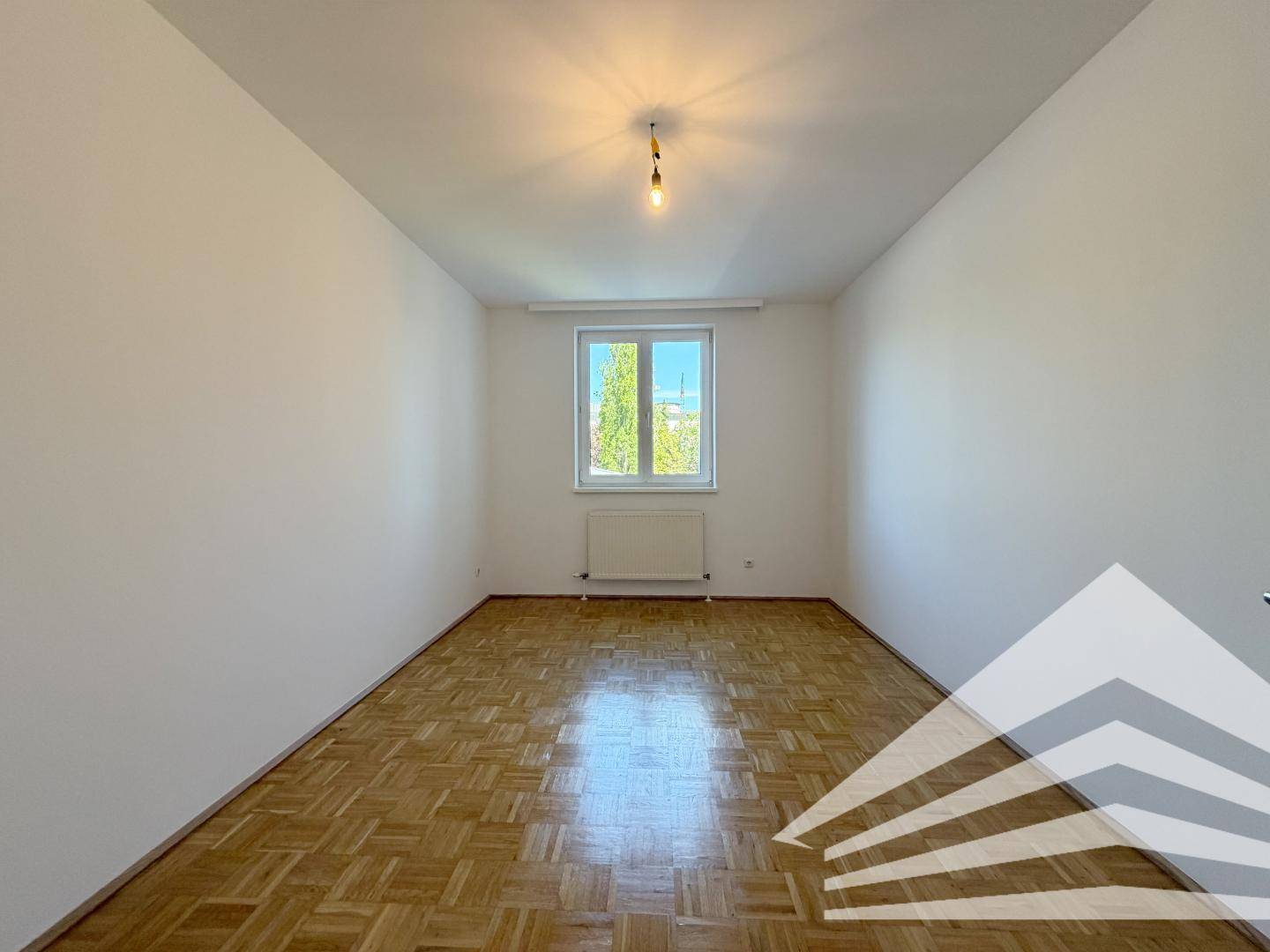 Helles Zimmer mit Parkettboden und einem Fenster mit Blick ins Grüne.