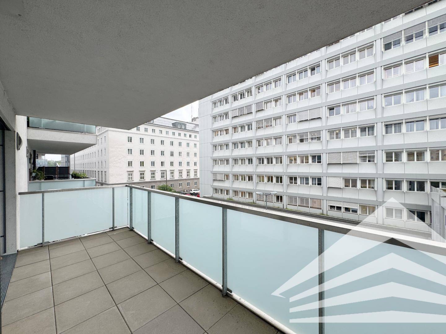 Balkon mit Fliesenboden und Milchglasgeländer, umgeben von städtischer Architektur.