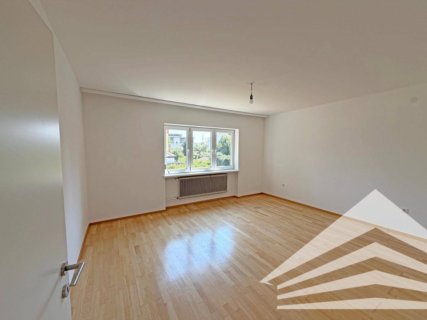 Helles, leeres Zimmer mit hellem Holzparkettboden und einem großen Fenster mit Blick ins Grüne.