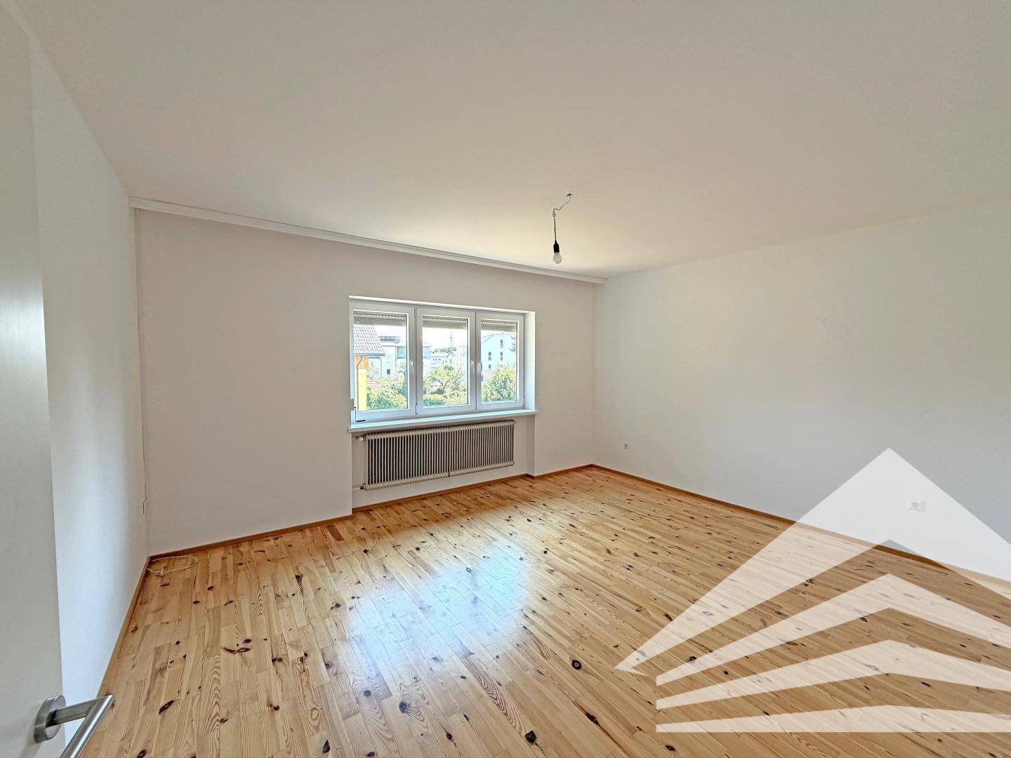 Helles, leeres Zimmer mit hellem Holzparkettboden und einem großen Fenster mit Blick ins Grüne.