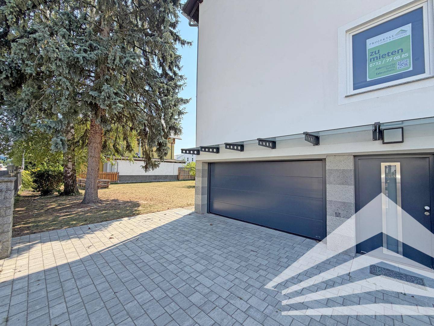 Moderne Hausfassade mit integrierter Garage, gepflasterter Einfahrt und einem großen Nadelbaum im Vorgarten.