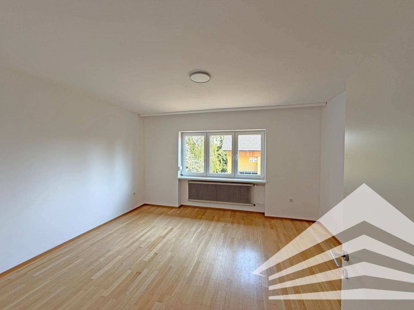 Helles Zimmer mit Holzparkettboden und einem Fenster, das den Blick auf die Umgebung freigibt.