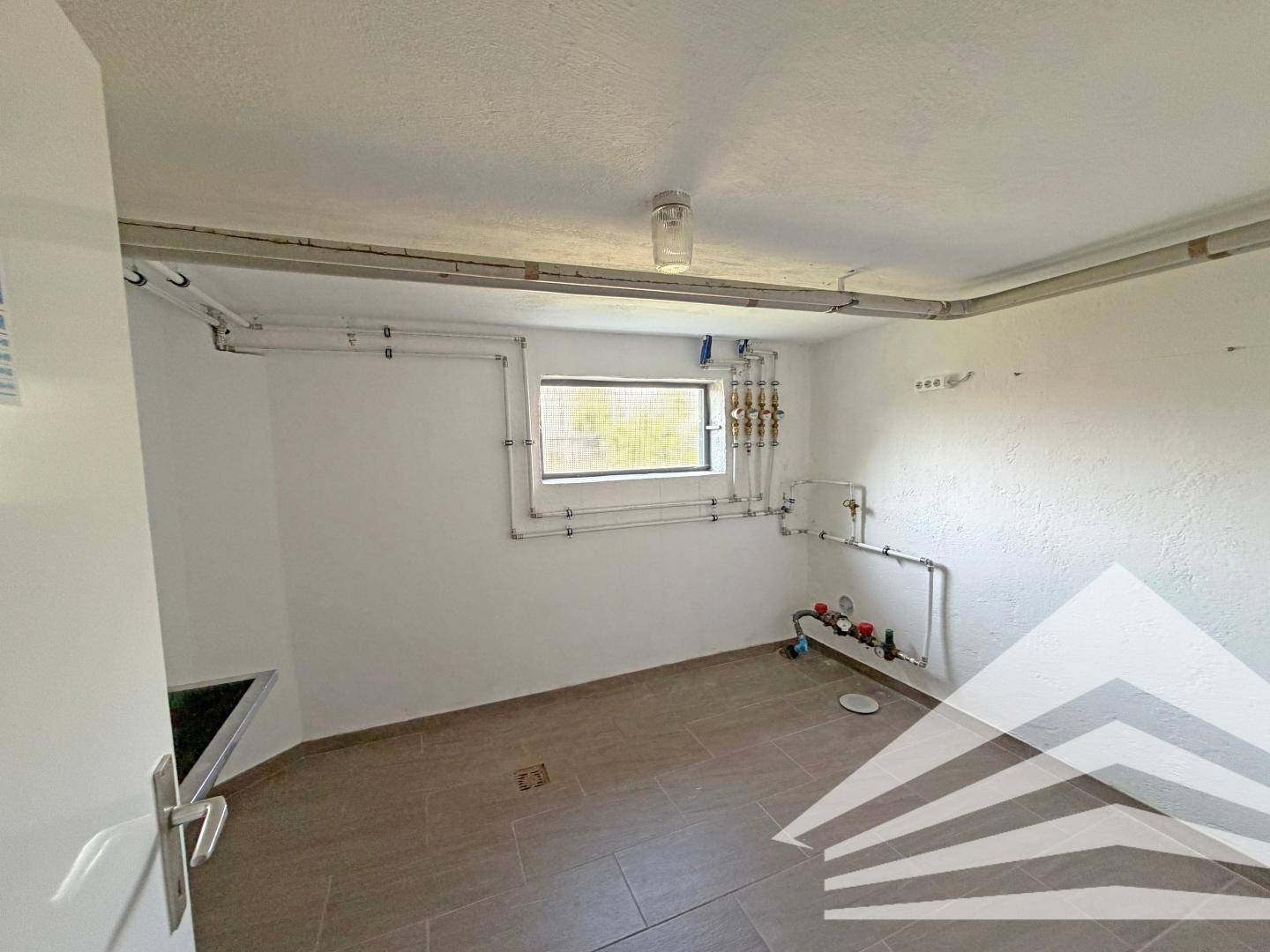 Funktionaler Hauswirtschaftsraum im Keller mit Fliesenboden, Fenster und sichtbaren Wasserleitungen an der Wand.