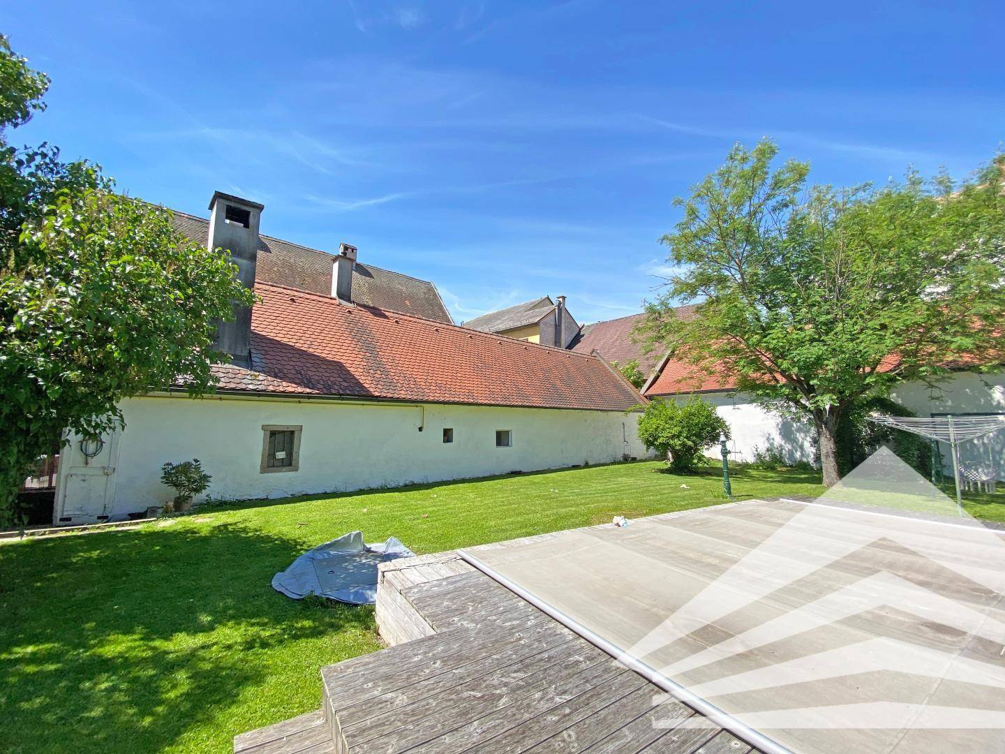 Gartenbereich mit Poolabdeckung und Rasenfläche, umgeben von alten Gebäuden unter blauem Himmel.