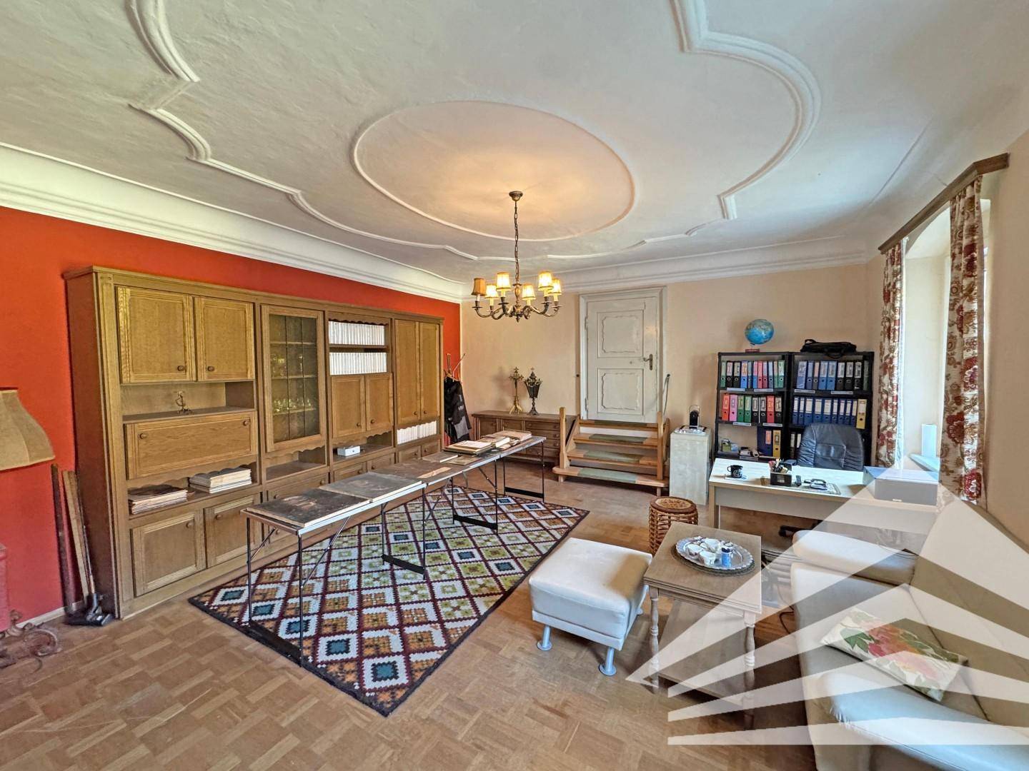 Großzügiges Wohnzimmer mit Parkettboden, roter Akzentwand und dekorativer Stuckdecke.