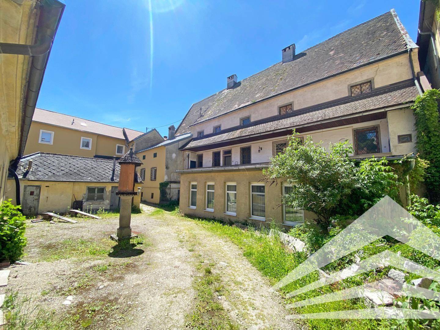 Hinterhof mit unbefestigtem Weg und alten Gebäuden, umgeben von üppigem Grün.