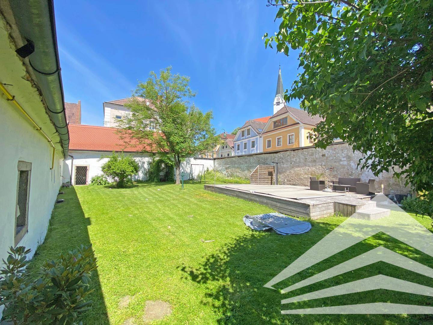 Großer grüner Garten mit Rasenfläche, Bäumen und einer Terrasse im Hintergrund, umgeben von historischen Gebäuden.