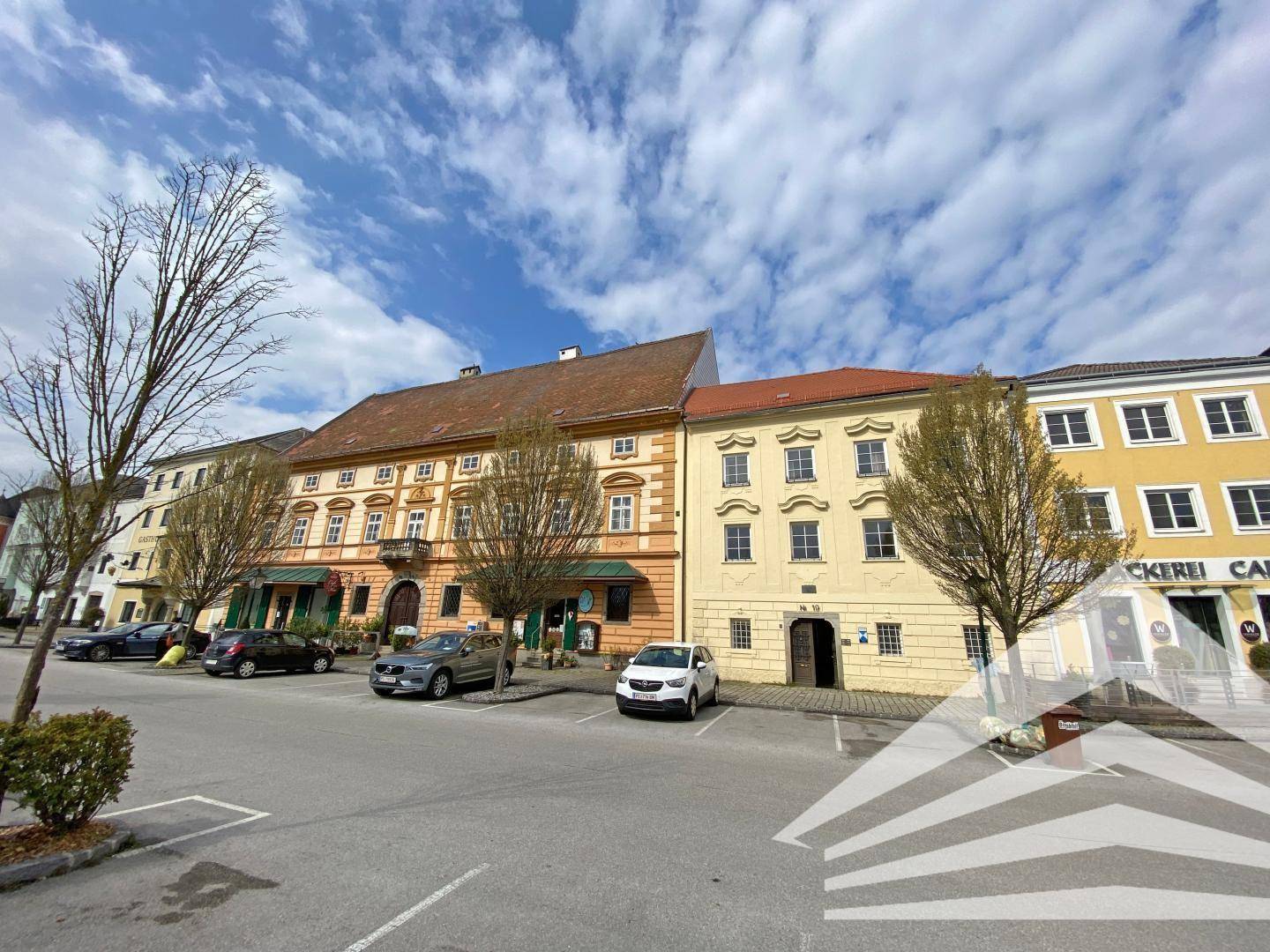 Historische Gebäudezeile an einem sonnigen Platz mit parkenden Autos und Bäumen.