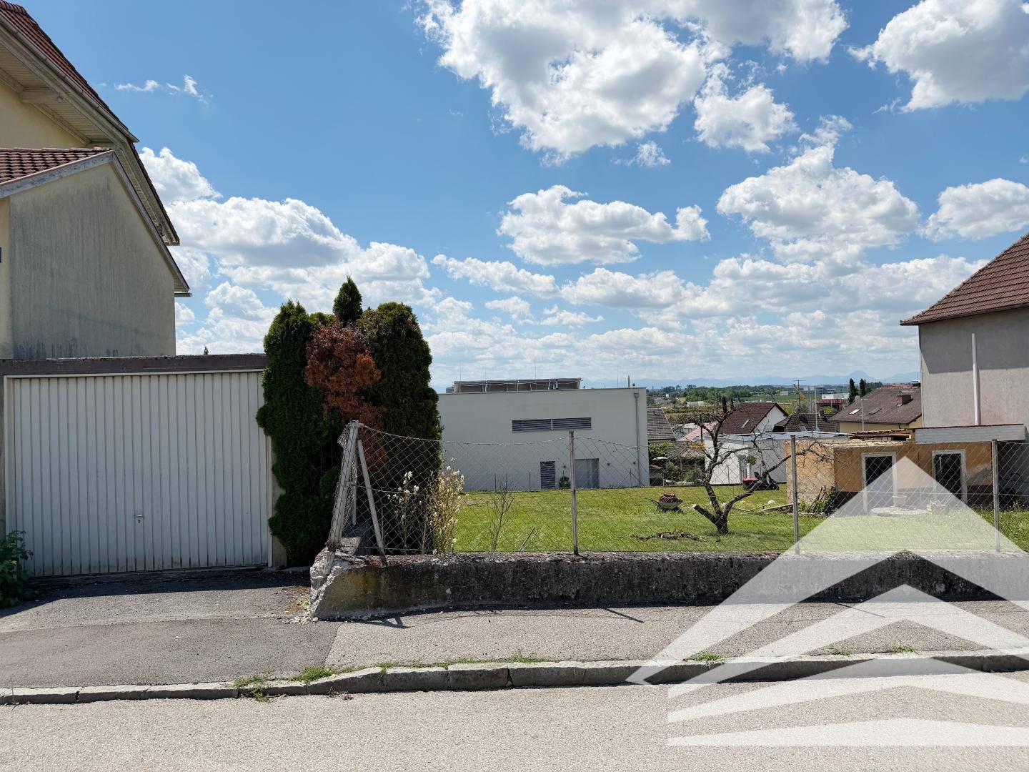 Ansicht einer Straße mit einer Garage und einem dahinterliegenden Grundstück unter blauem Himmel.