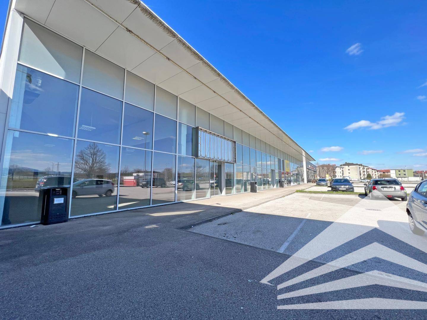 Moderne Geschäftsfassade mit großen Glasflächen und einem weitläufigen Parkplatz unter blauem Himmel.