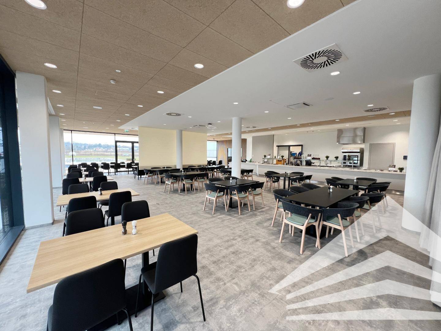 Geräumige Kantine mit vielen Tischen und Stühlen, großer Fensterfront und moderner Essensausgabe.