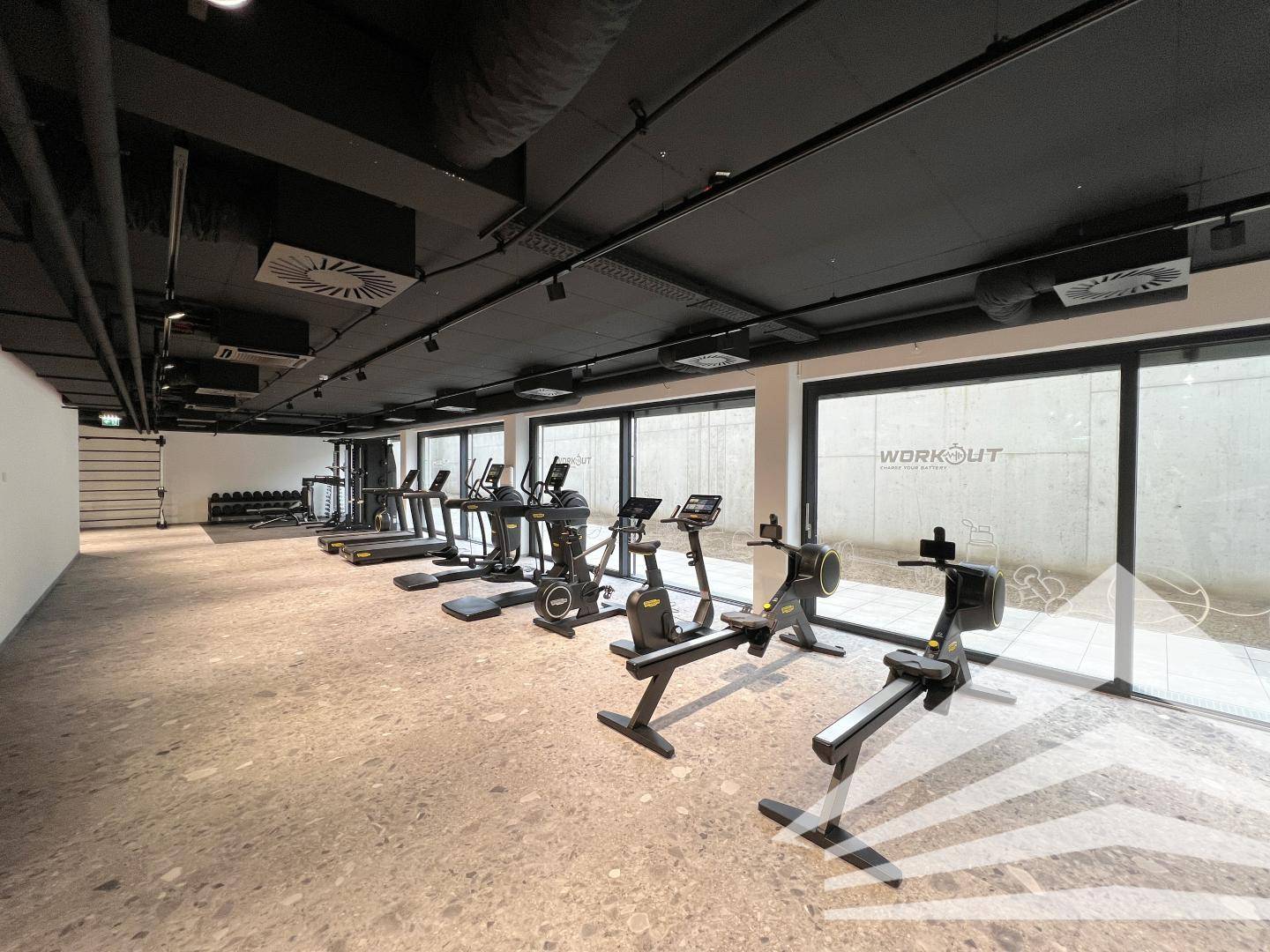 Moderner Fitnessraum mit verschiedenen Trainingsgeräten, großen Fenstern und industriellem Decken-Design.