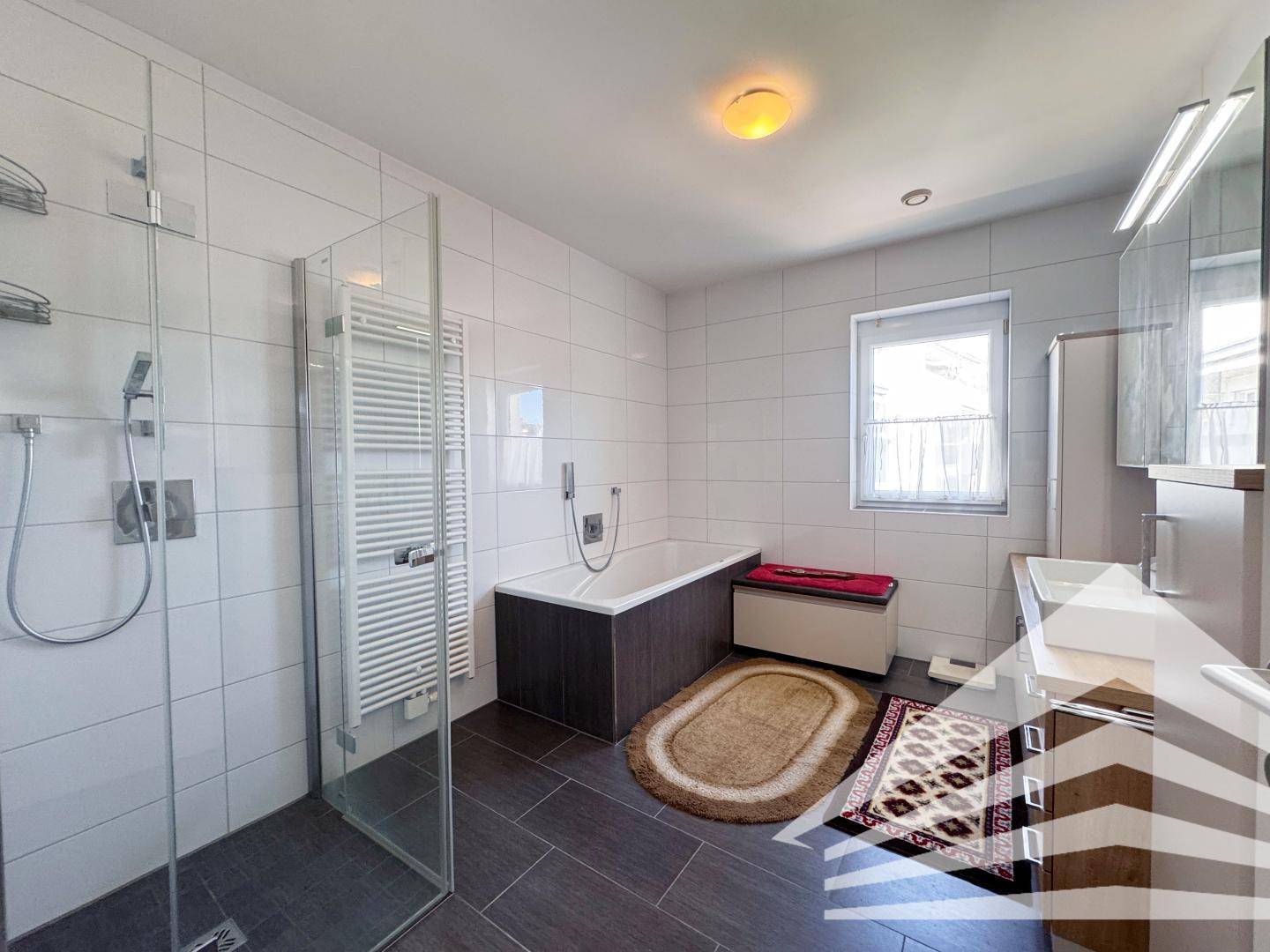 Modernes Badezimmer mit Dusche, Badewanne und Doppelwaschbecken, hell gefliest und mit Fenster.