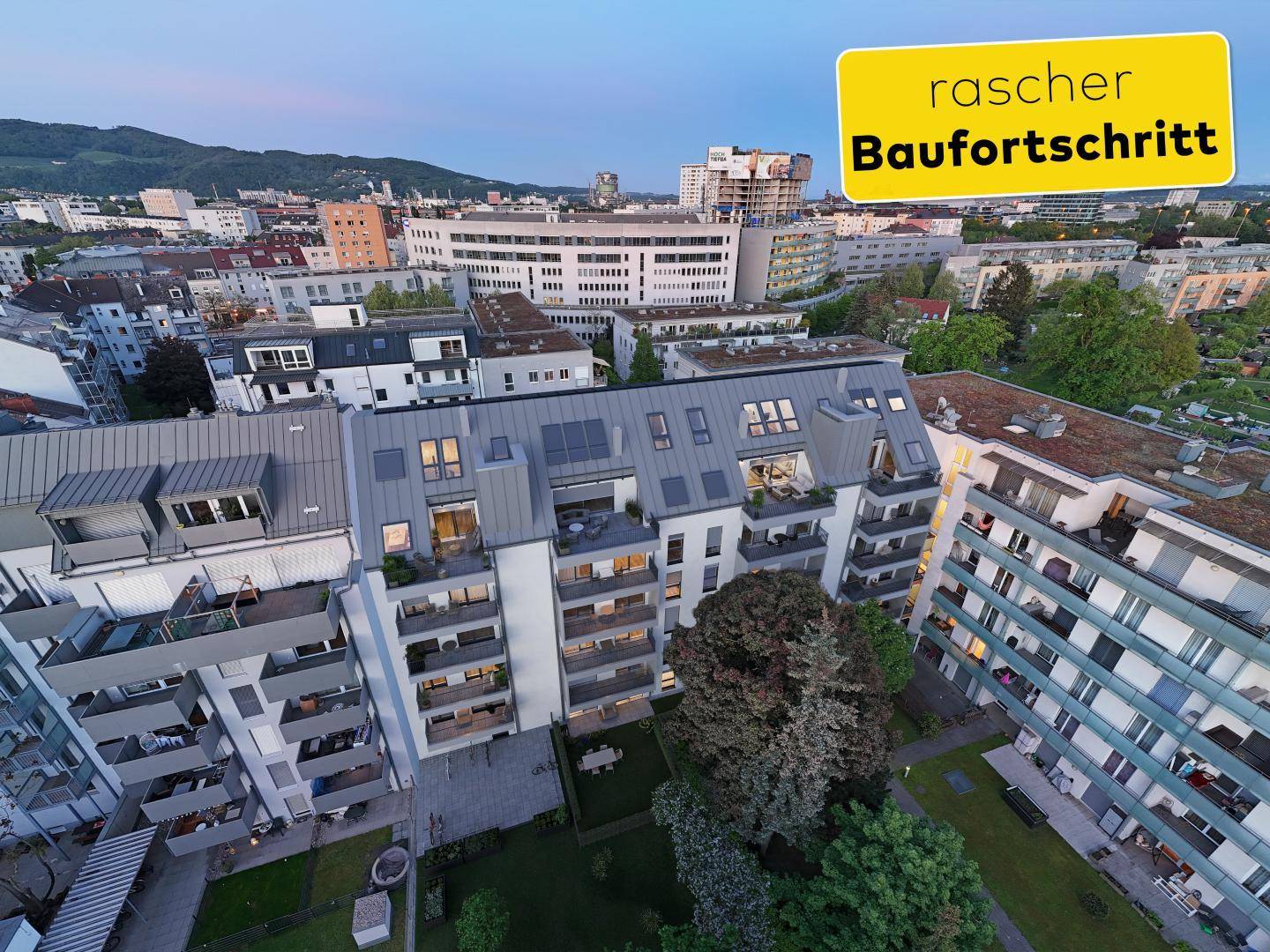 Luftaufnahme einer modernen Wohnanlage bei Dämmerung mit begrüntem Innenhof und Stadtblick.