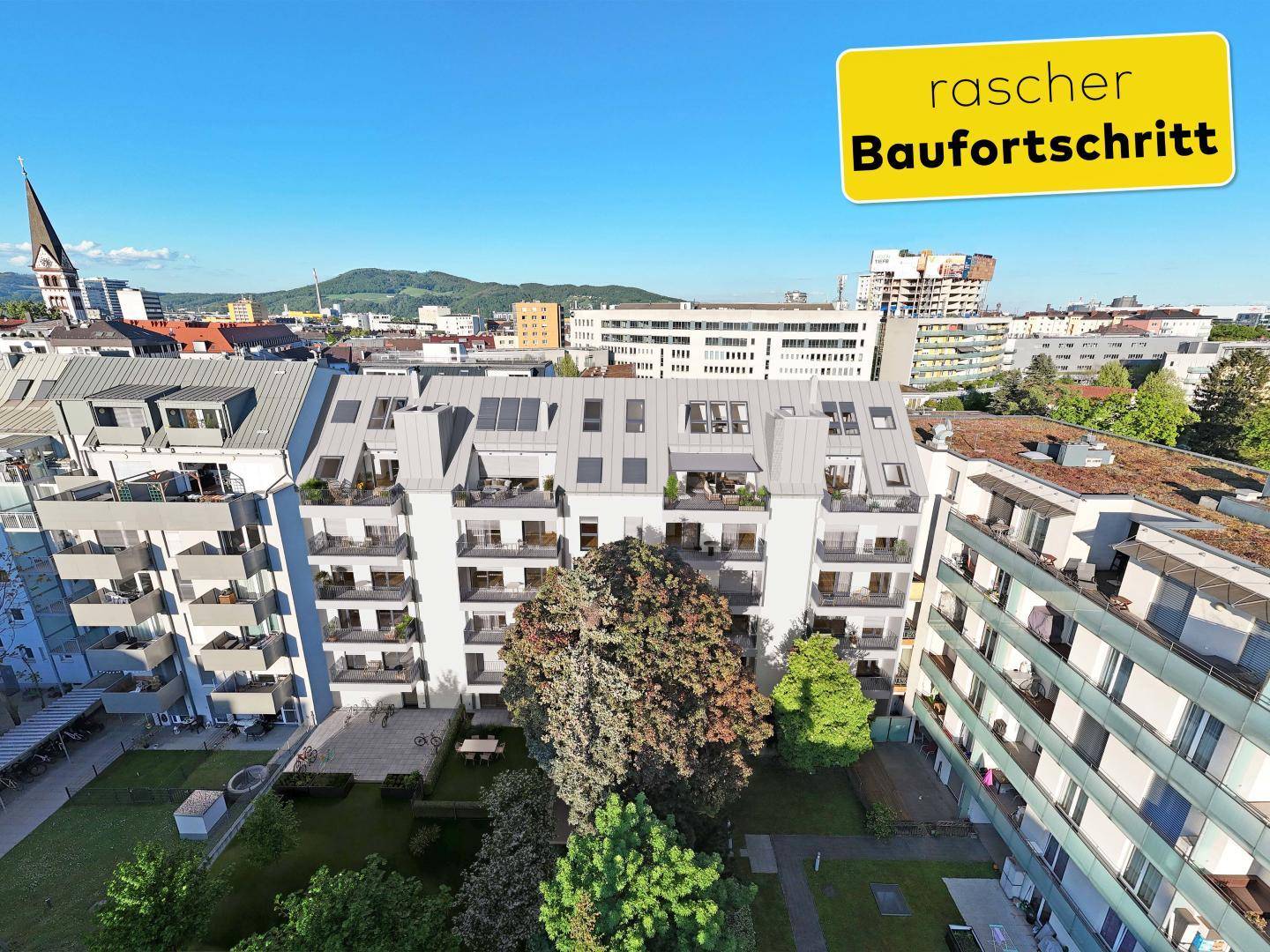 Luftaufnahme des modernen Wohngebäudes mit umliegender Stadtlandschaft und Kirchturm im Hintergrund.