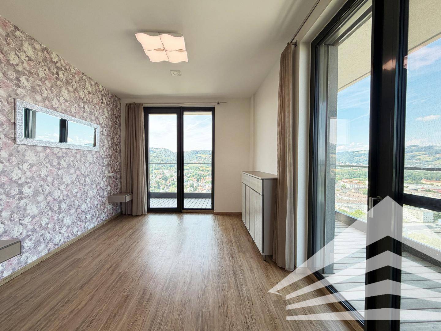 Helles Zimmer mit dekorativer Tapete und großen Fenstern, die einen weiten Ausblick bieten.