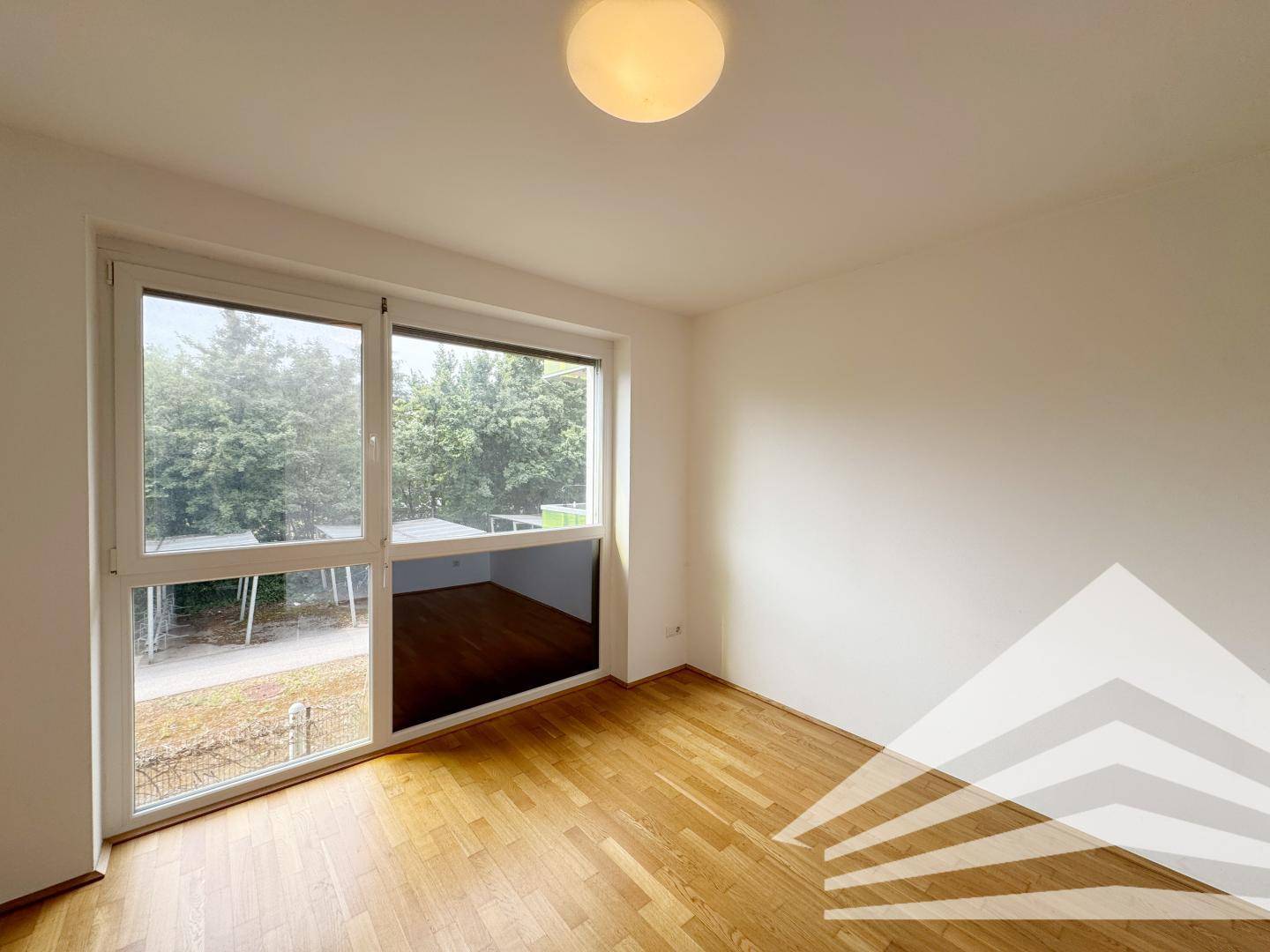 Helles Zimmer mit Holzboden und großen Fenstern, die viel Tageslicht hereinlassen.