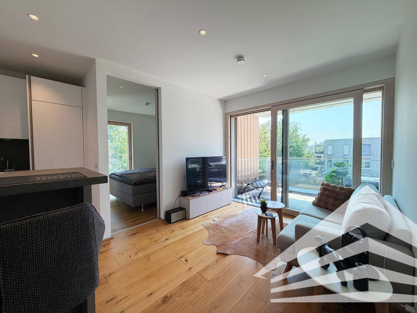 Helles Wohnzimmer mit Holzboden, Zugang zum Balkon und Blick ins Schlafzimmer.