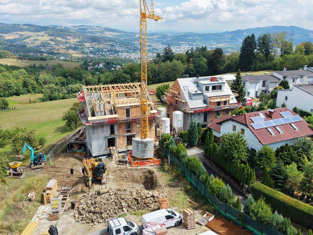Baustelle mit zwei im Bau befindlichen Häusern und einem Kran, umgeben von grüner Landschaft und weiteren Wohnhäusern.