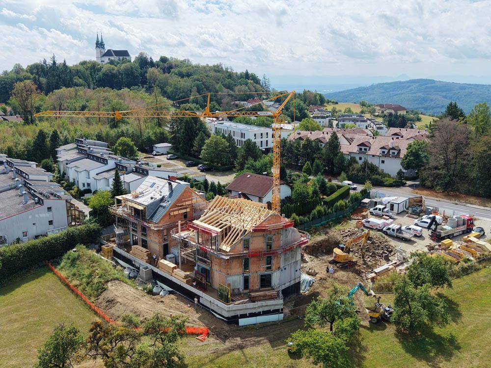 Baustelle mit zwei im Bau befindlichen Häusern und einem Kran, umgeben von grüner Landschaft und einem Hügel mit Kirche.