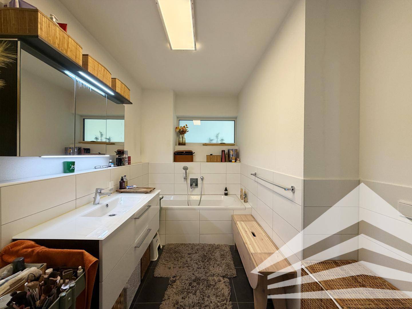 Helles Badezimmer mit Badewanne, Waschtisch und großem Spiegel, teilweise gefliest.