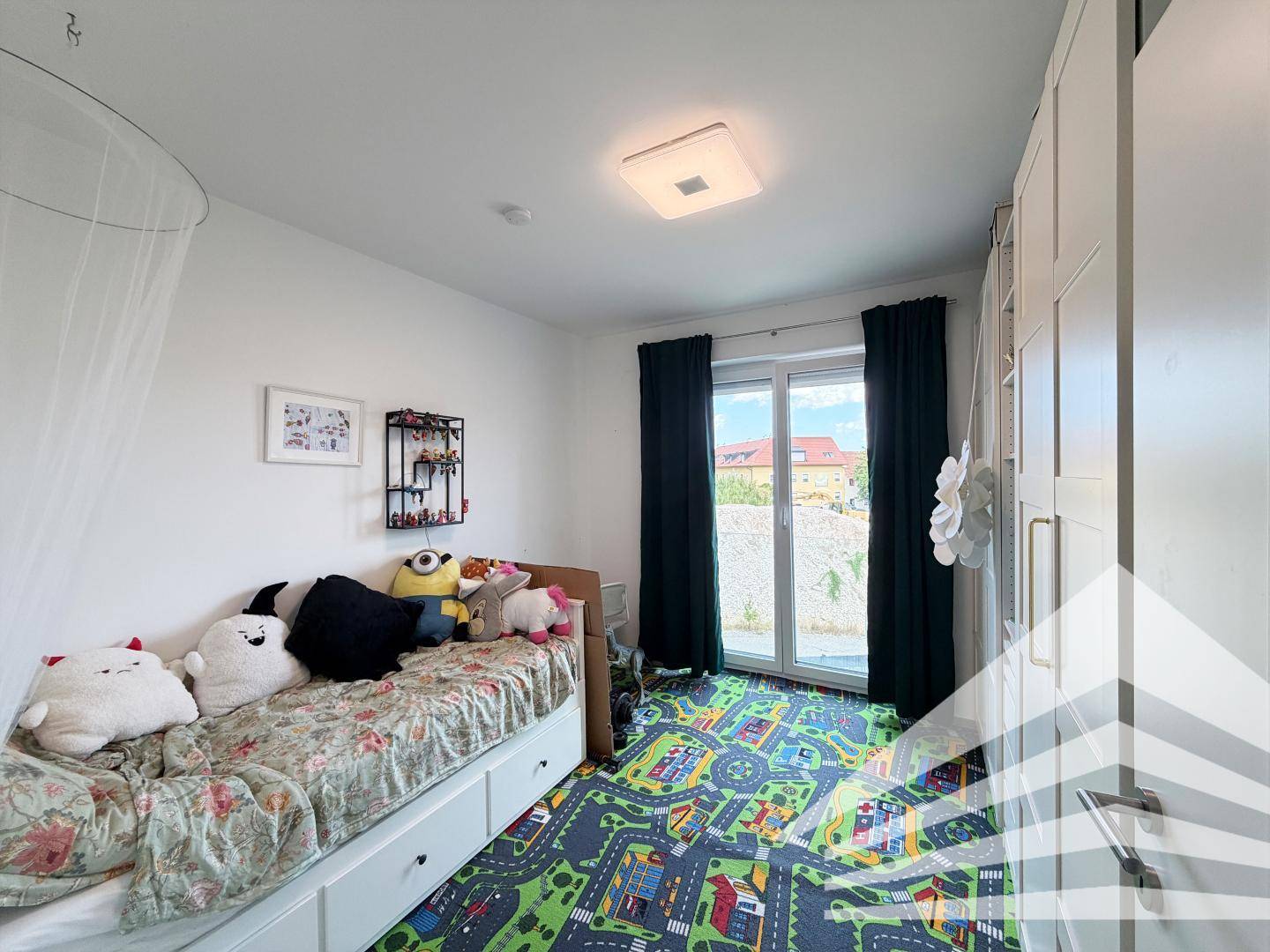 Kinderzimmer mit Einzelbett, Spielteppich und großem Fenster für viel Tageslicht.