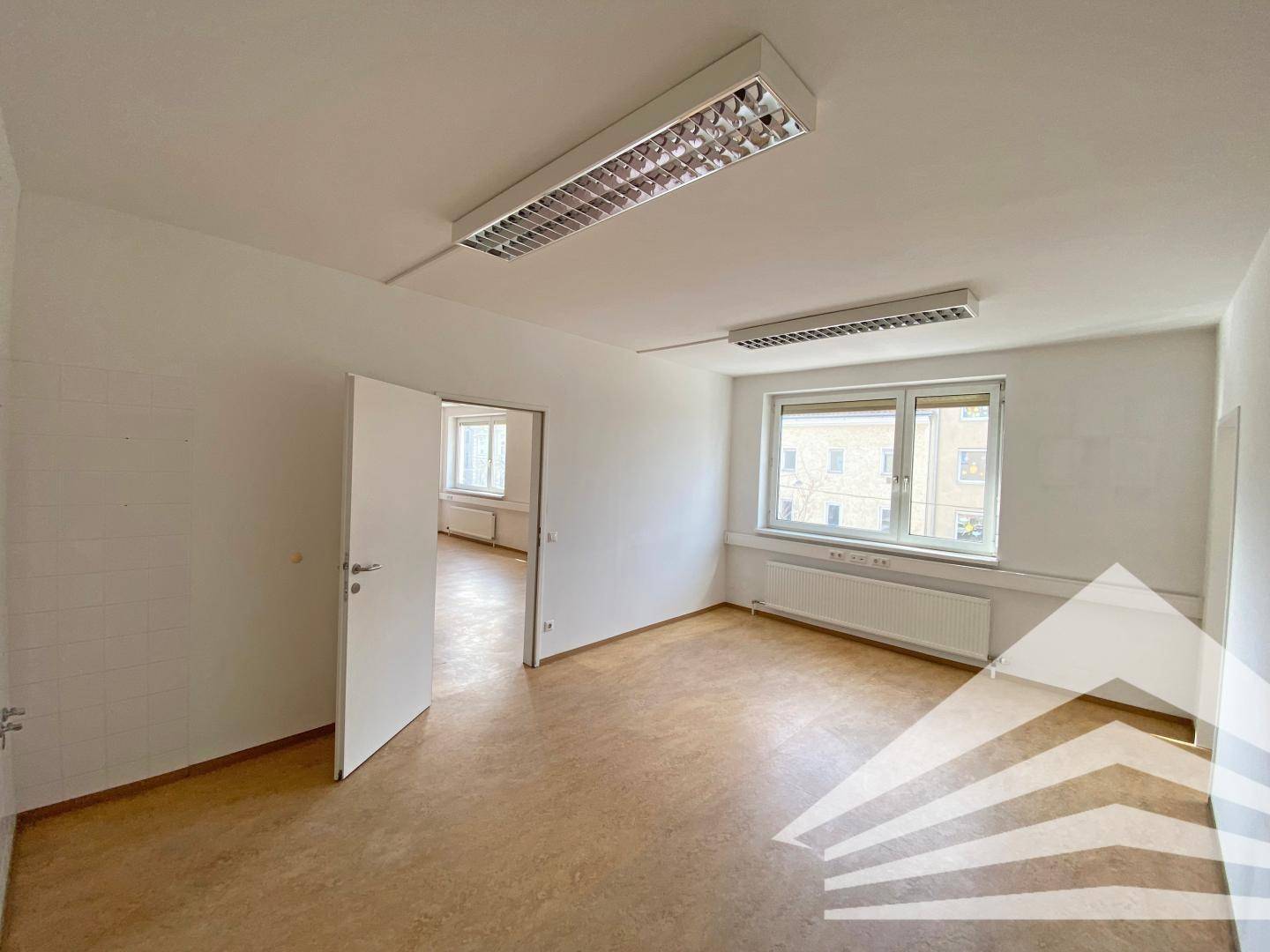Leerer Büroraum mit offener Tür zu einem weiteren Zimmer, ausgestattet mit großen Fenstern.