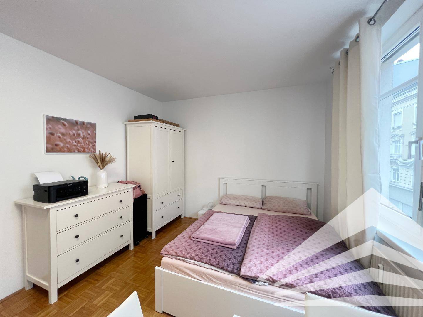 Schlafzimmer mit Doppelbett, Kommode und Kleiderschrank, hell beleuchtet durch das große Fenster.