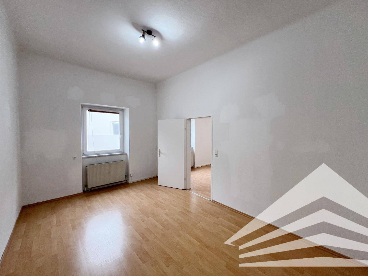 Helles Zimmer mit Parkettboden, Fenster und Zugang zu einem weiteren Raum.