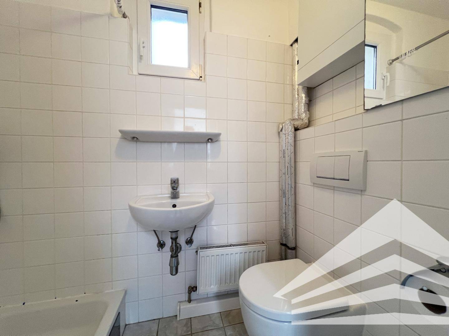 Einfaches Badezimmer mit weißen Fliesen, Waschbecken, Toilette und Fenster.