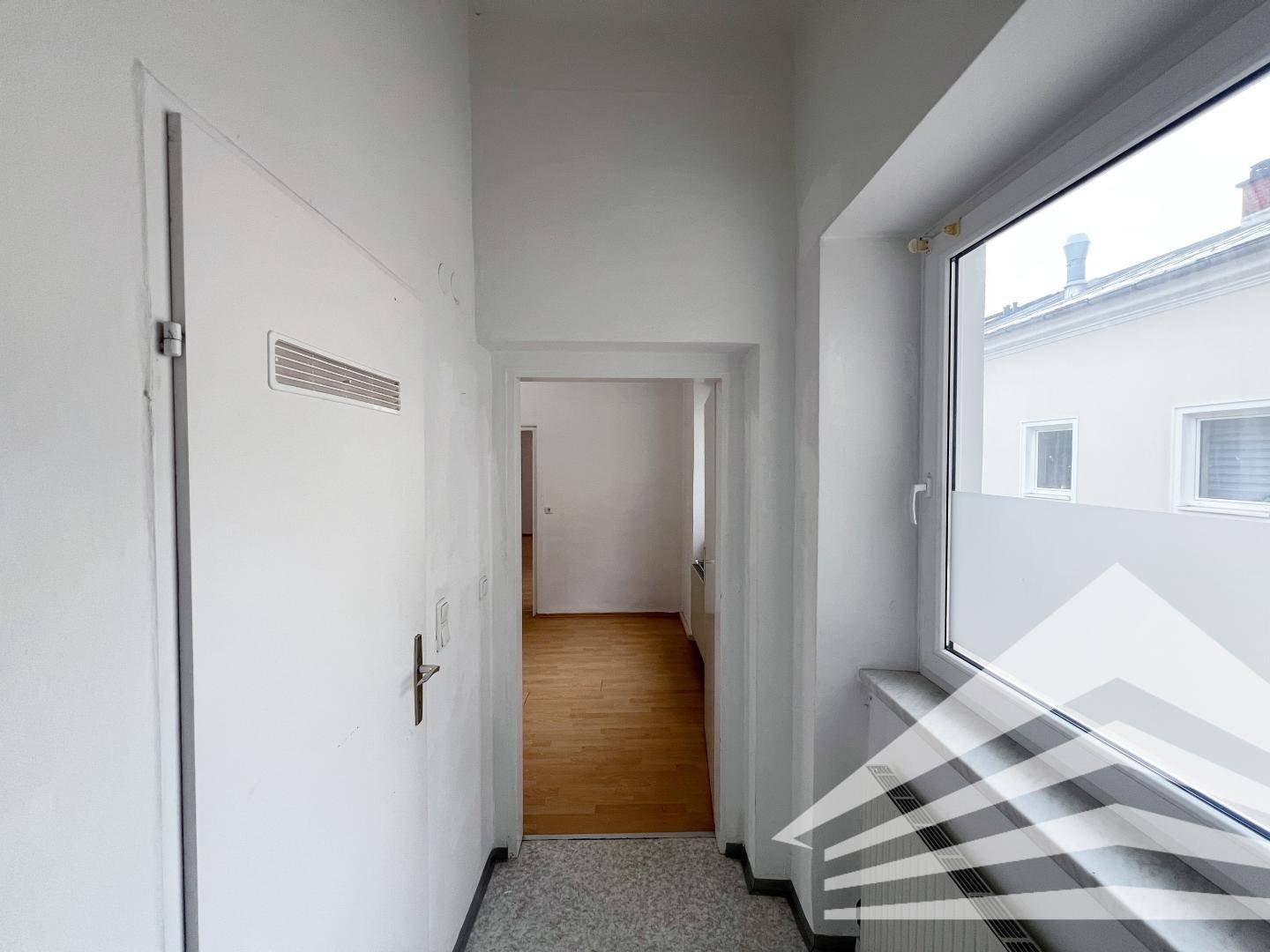 Schmaler Flur mit Fenster und Blick in ein angrenzendes Zimmer mit Parkettboden.