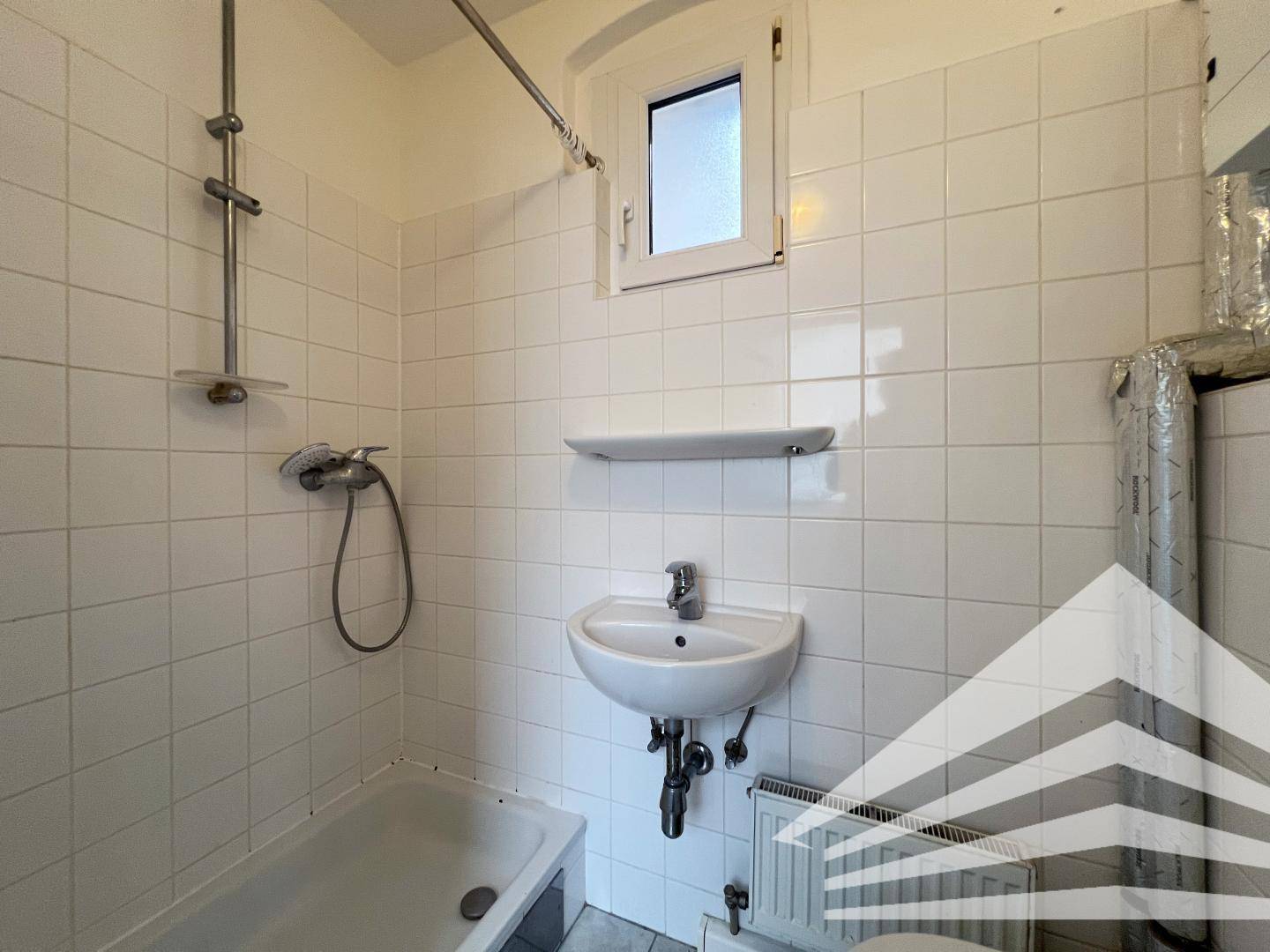 Funktionales Badezimmer mit weißen Fliesen, Waschbecken, Duschbereich und Fenster.