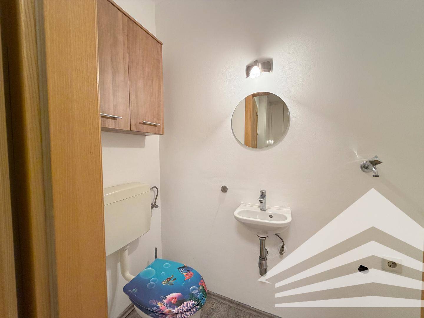 Gäste-WC mit kleinem Waschbecken, Spiegel und farbenfrohem Toilettendeckel.