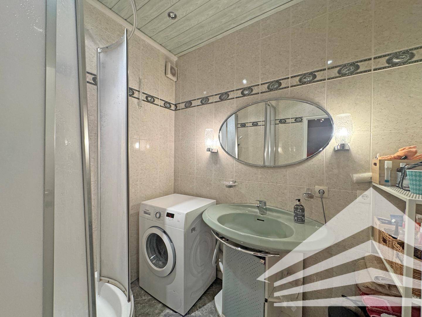 Gefliestes Badezimmer mit Dusche, Waschbecken und Waschmaschine, sowie Holzdecke.