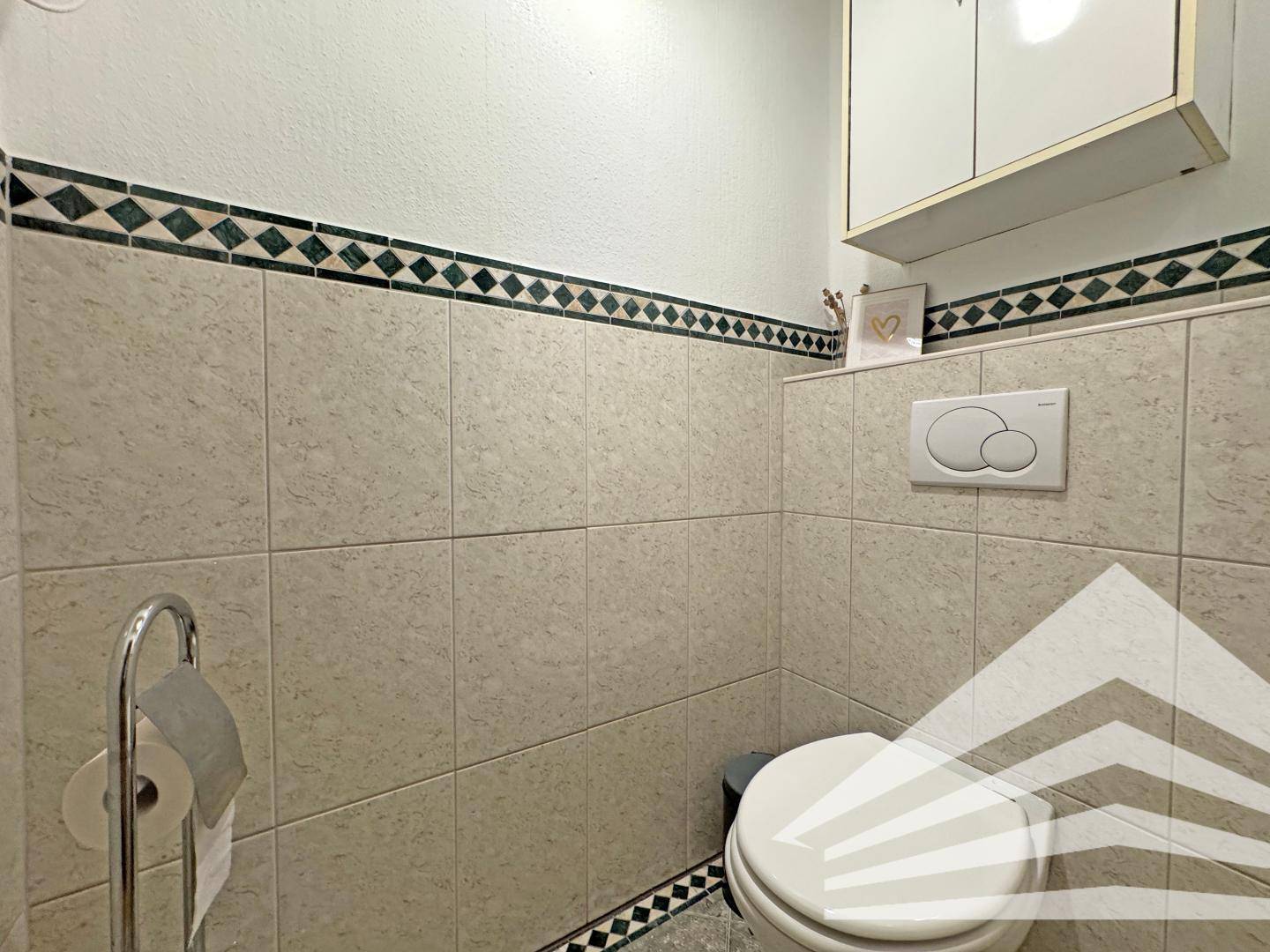 Separate Toilette mit hellen Fliesen und dekorativer Bordüre, sowie Wand-WC.