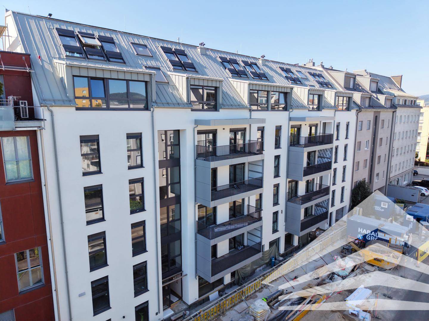Neubau-Fassade mit Balkonen und Dachfenstern, umgeben von städtischer Bebauung.