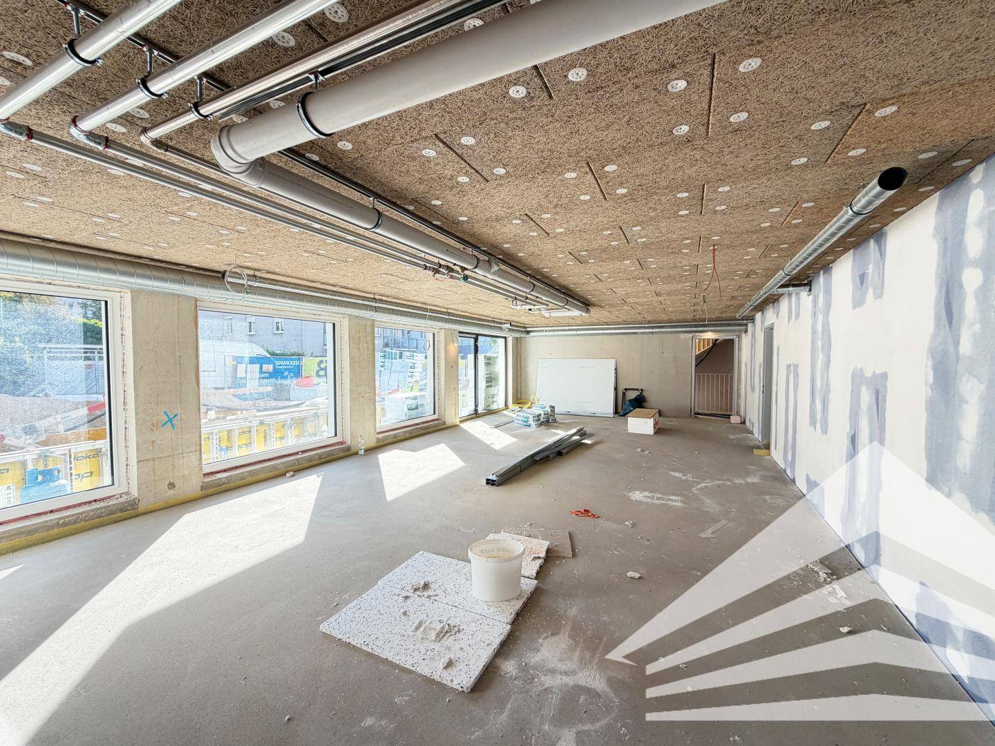Unfertiger Innenraum mit sichtbaren Rohren an der Decke und großen Fenstern im Rohbau.