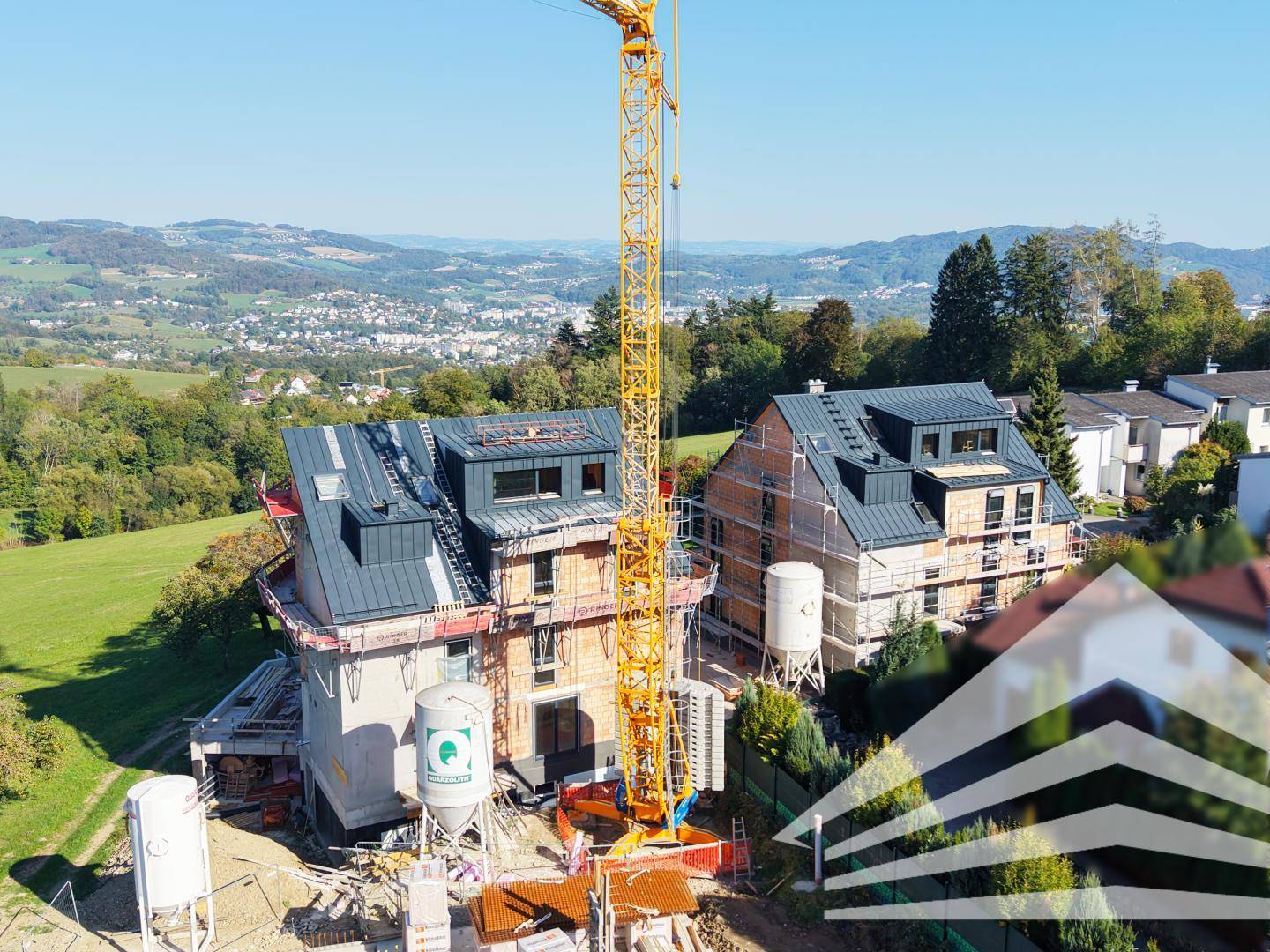 Baustelle mit zwei im Bau befindlichen Gebäuden und einem hohen Kran vor einer hügeligen Landschaft.
